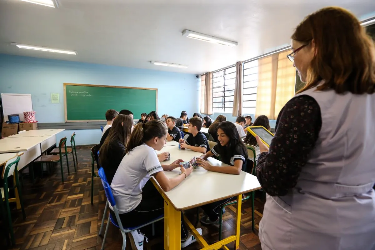 Na imagem, uma professora de costas dá aula em uma sala com alunos sentados, que dividem grandes mesas.