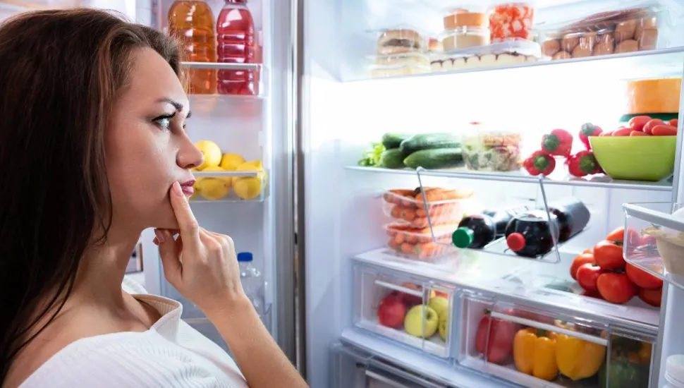 Imagem mostra uma mulher olhando com dúvida para uma geladeira aberta. TEm muitos produtos dentro com tomate, pimentão, bebidas.