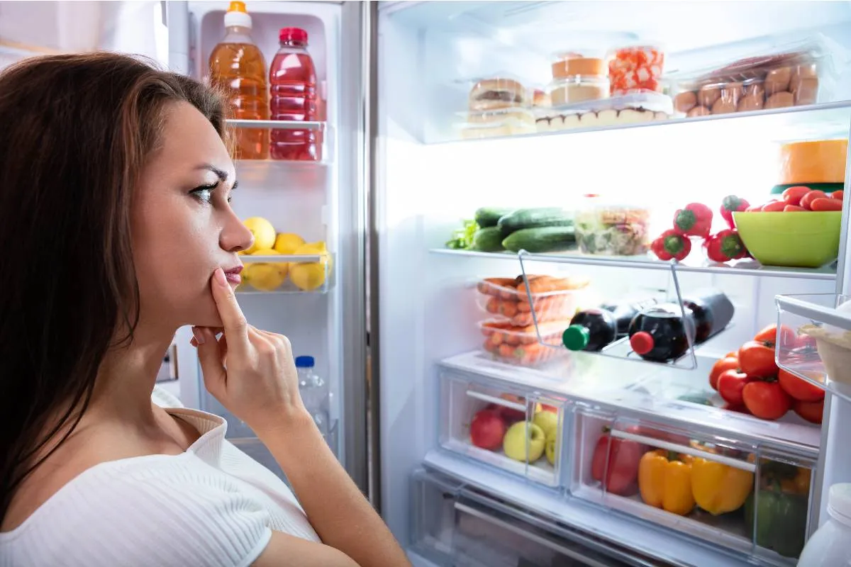 Imagem mostra uma mulher olhando com dúvida para uma geladeira aberta. TEm muitos produtos dentro com tomate, pimentão, bebidas.