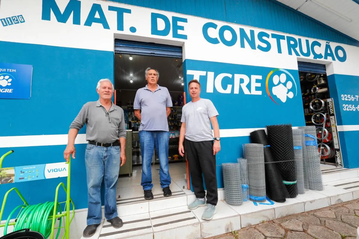 Imagem mostra três homens em um balcão de um material de construção com ferramentas em uma bancada.