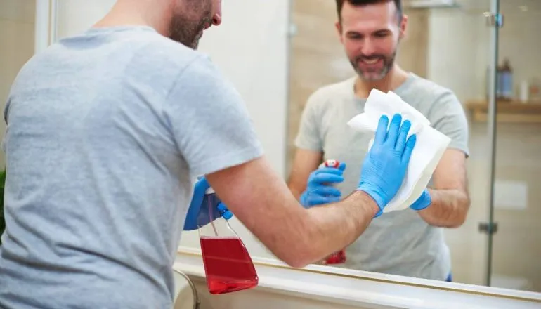 Imagem mostra um homem com uma luva azul e um borrifador com líquido vermelho limpando um espelho. 