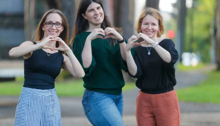 Imagem mostra três mulheres sorrindo e fazendo coração com as mãos.