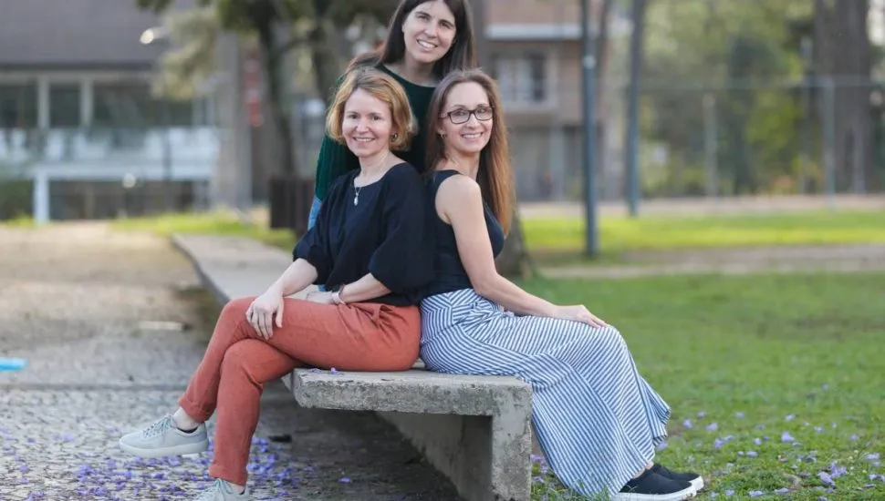 Imagem mostra três mulheres sentadas em um banco sorrindo.