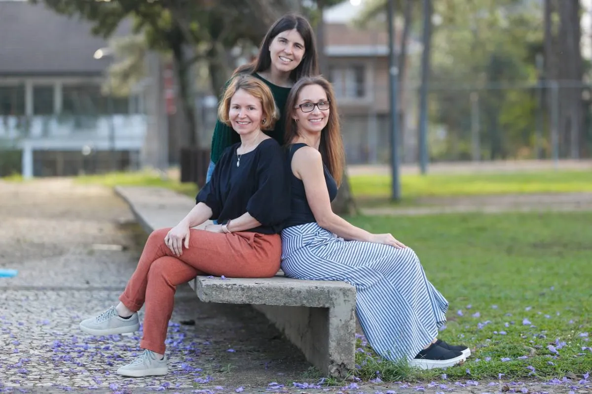 Imagem mostra três mulheres sentadas em um banco sorrindo.