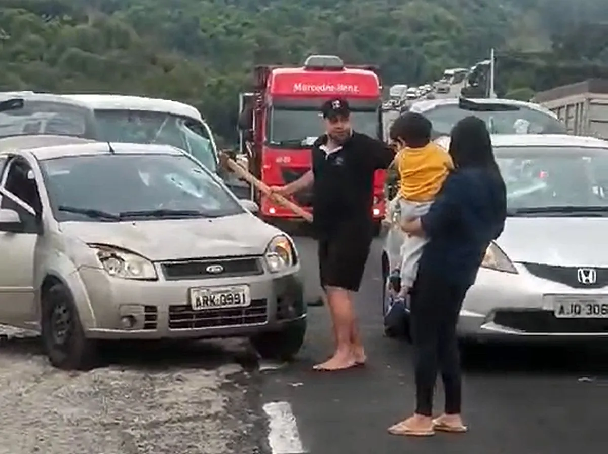 homem segura um machado e é observado por uma mulher com criança no colo. Dois carros aparecem na imagem, um deles destruído pela ação do homem com o machado