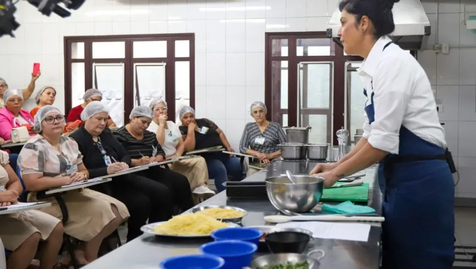 Na imagem, aula da chef Manu Buffara para merendeiras, que assistem sentadas na plateia.