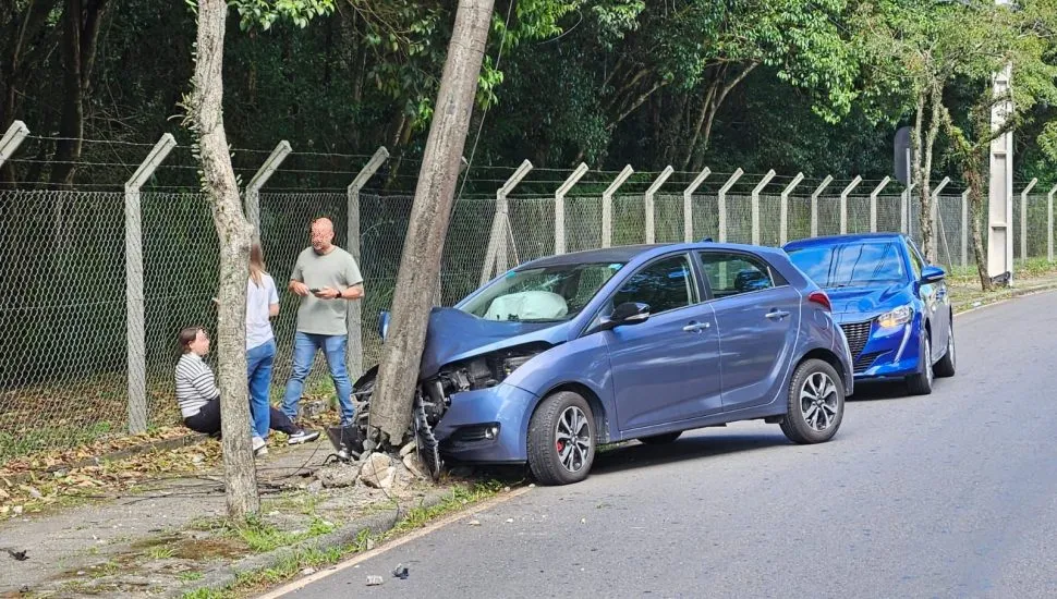 imagme mostra carro batido em poste. A motorista está sentada no chão e outras duas pessoas, um homem e uma mulher, lhe prestam assistência