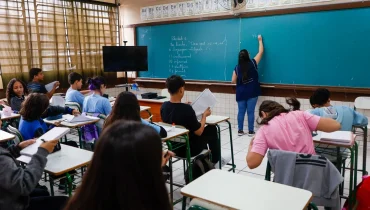 Na imagem, uma professora escreve no quadro. Alunos estão na sala, observando aula sentados.