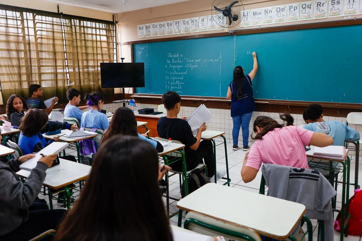 Na imagem, uma professora escreve no quadro. Alunos estão na sala, observando aula sentados.