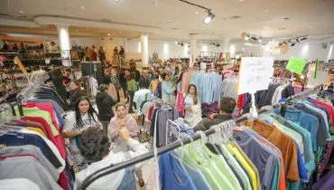 Na imagem, roupas em araras e clientes fazendo compras no Bazar Moda do Bem