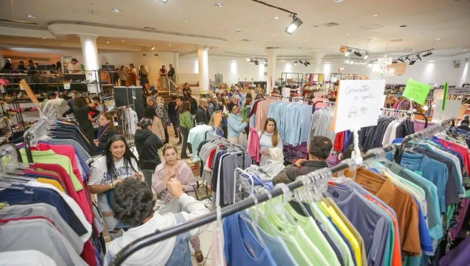 Na imagem, roupas em araras e clientes fazendo compras no Bazar Moda do Bem