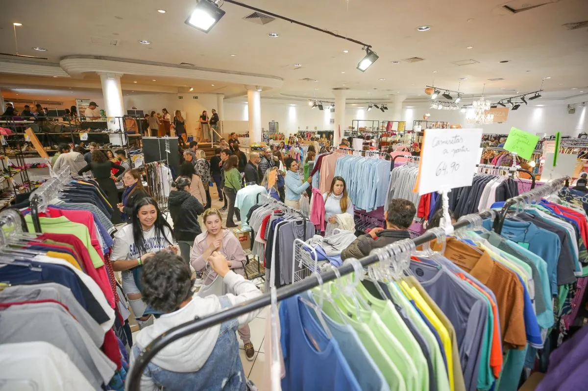 Na imagem, roupas em araras e clientes fazendo compras no Bazar Moda do Bem