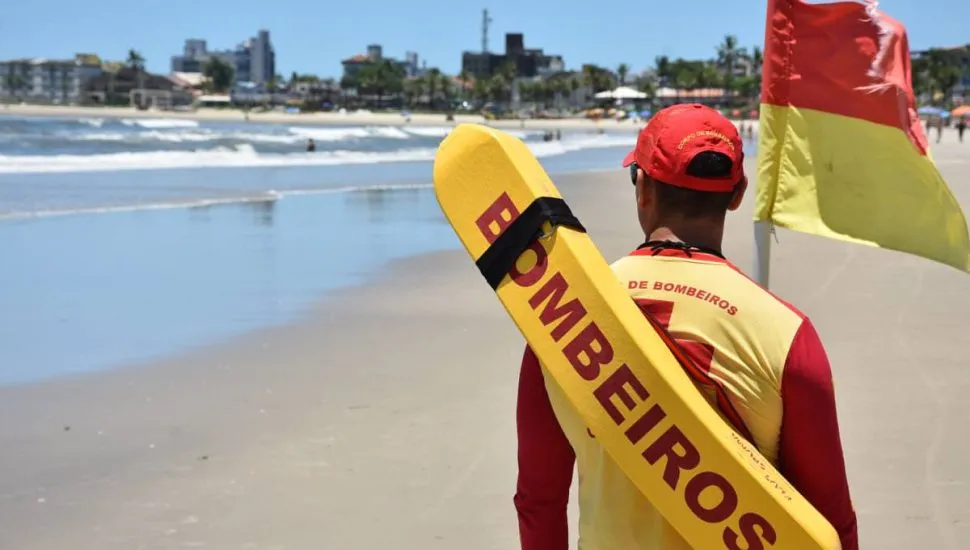 Imagem mostra um bombeiro em uma praia com uma boia de salvamento nas costas.