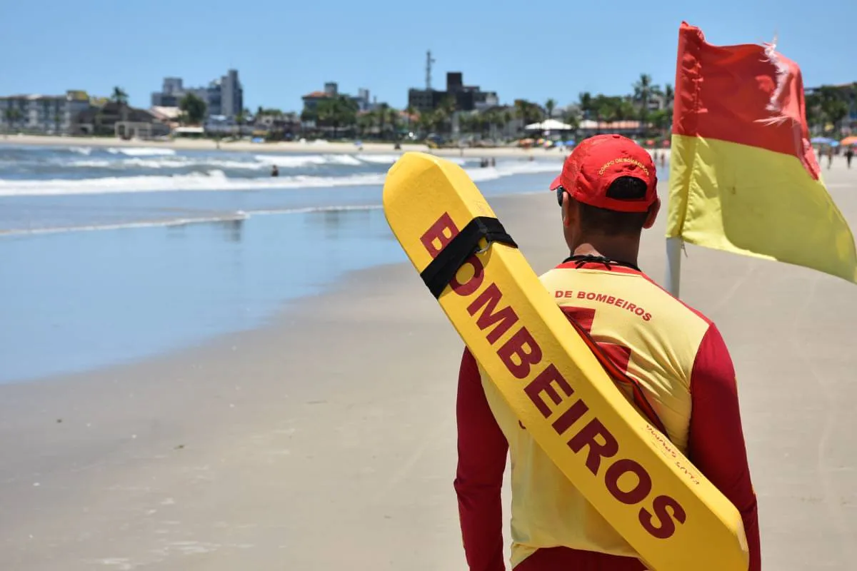 Imagem mostra um bombeiro em uma praia com uma boia de salvamento nas costas.