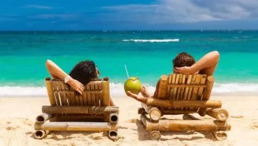 Na imagem, casal em cadeiras de praia, em frente ao mar, tomando água de coco