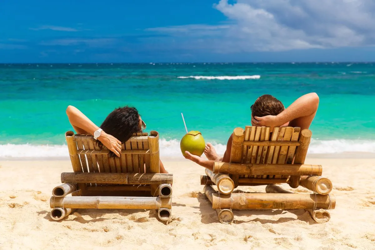 Na imagem, casal em cadeiras de praia, em frente ao mar, tomando água de coco