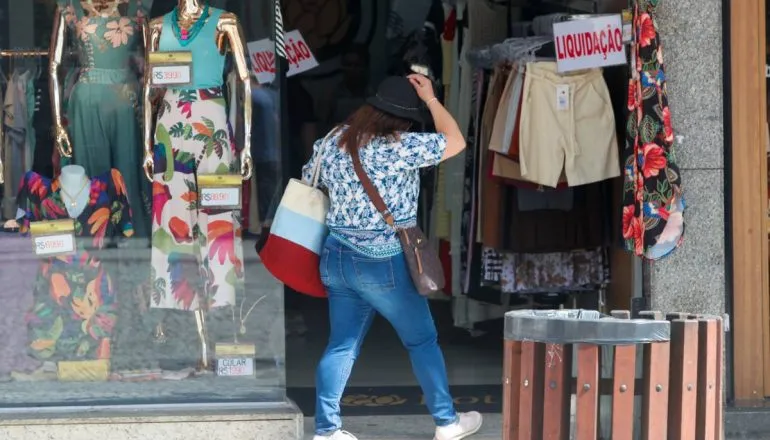 Imagem mostra uma mulher em frente a uma loja de roupa. Ela usa calça jeans, blusa azul e está segurando uma sacola com as cores vermelha, azul e branca.