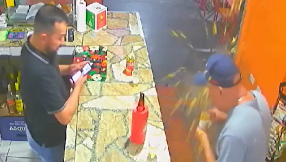 Imagem de câmera de segurança mostra um balcão de bar com um homem vestindo camiseta preta de um lado do balcão e, do outro, um homem de boné azul e camiseta azul com uma coxinha explosiva na mão.