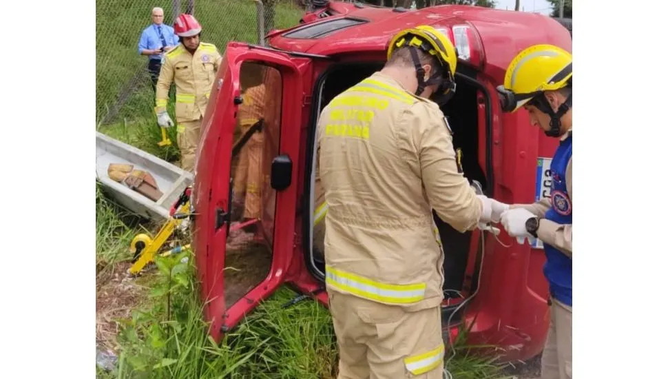 Imagem mostra um carro vermelho capotado com bombeiros ao lado.