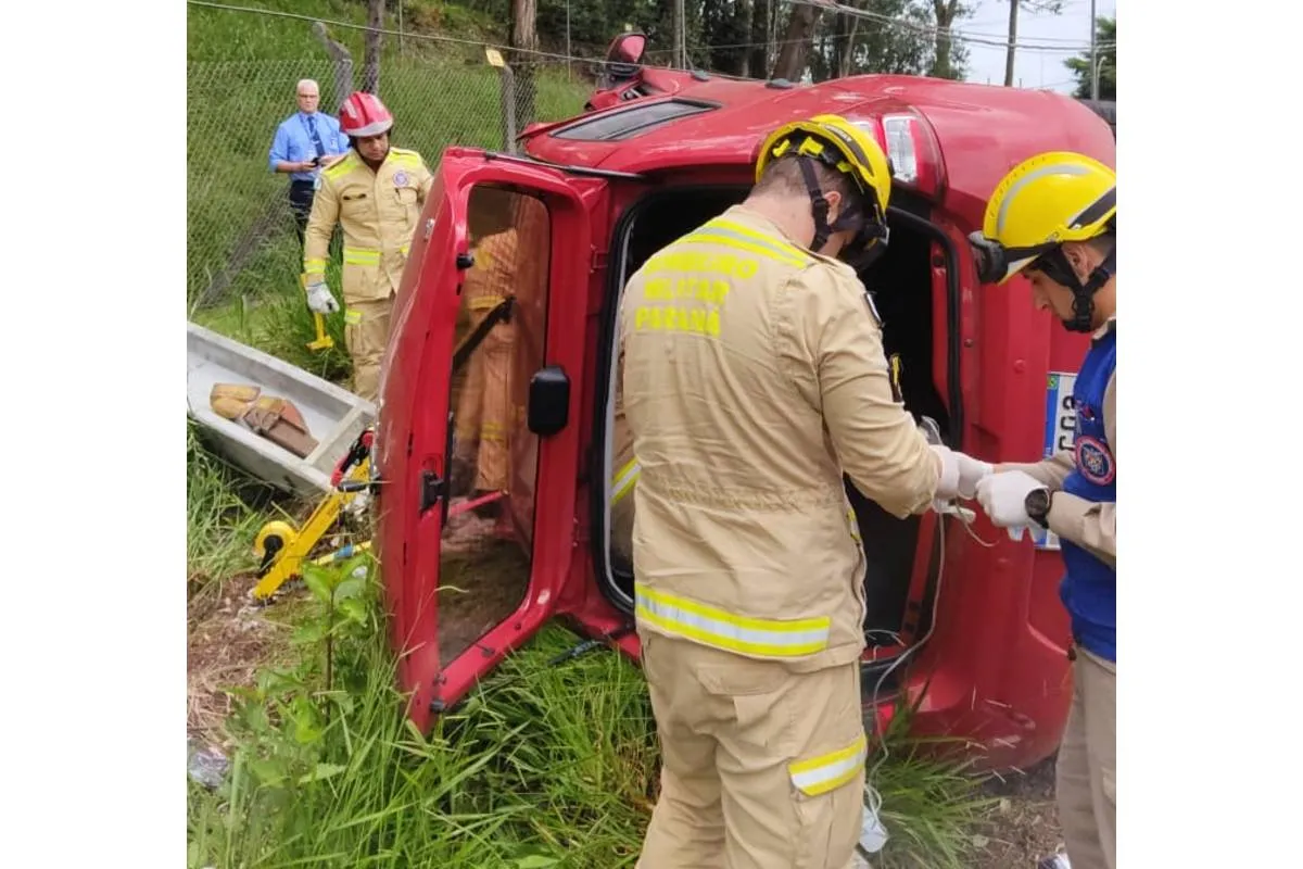 Imagem mostra um carro vermelho capotado com bombeiros ao lado.