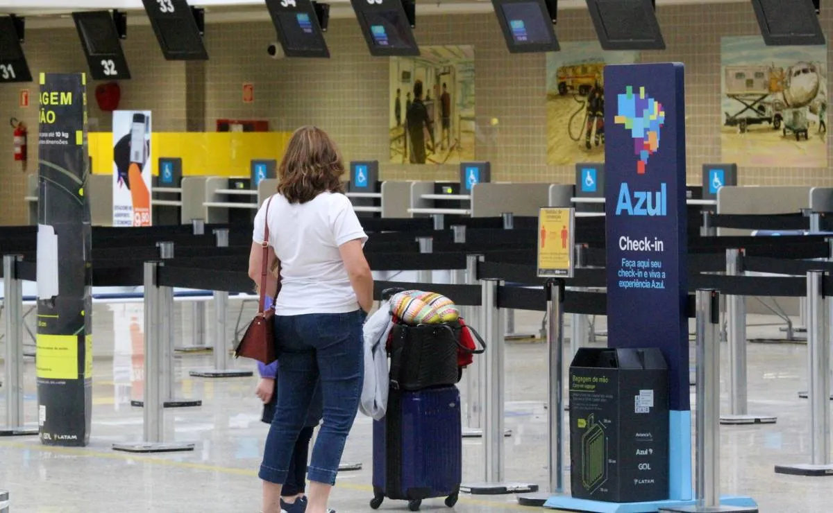 Imagem mostra uma passageira em um guichê de uma companhia aéra. Ela usa calça jeans e camiseta branca e está com malas de rodinha.