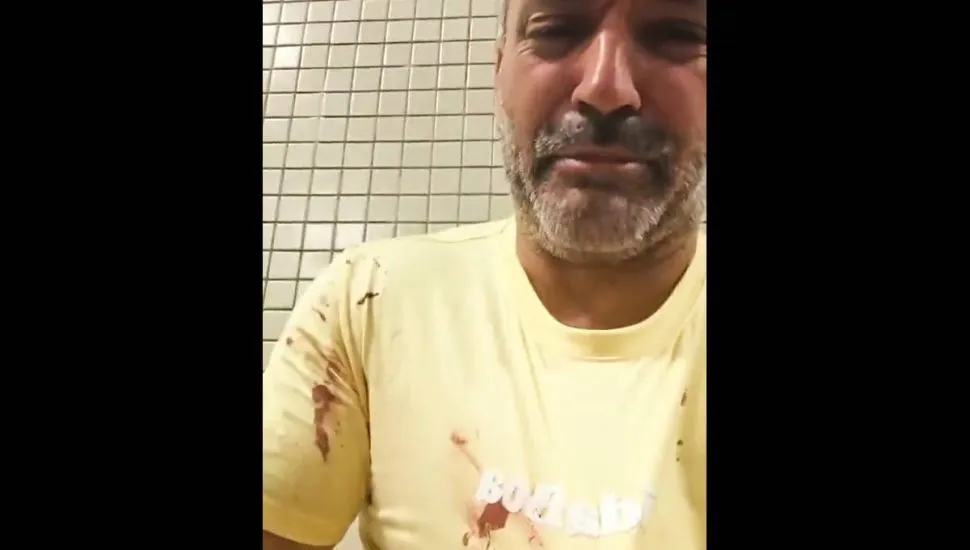 Imagem mostra um homem com camiseta amarela com manchas de sangue.