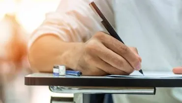 Imagem mostra uma pessoa em uma mesa usando uma caneta para escrever em um caderno.