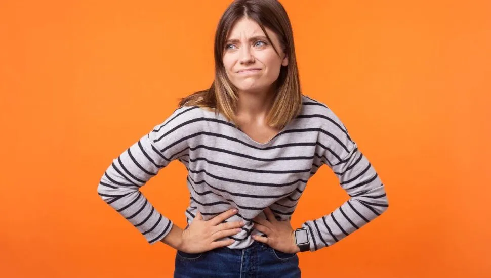 Imagem mostra uma mulher com dor de barriga, Ela está em um fundo laranja usando blusa listrada e calça preta.