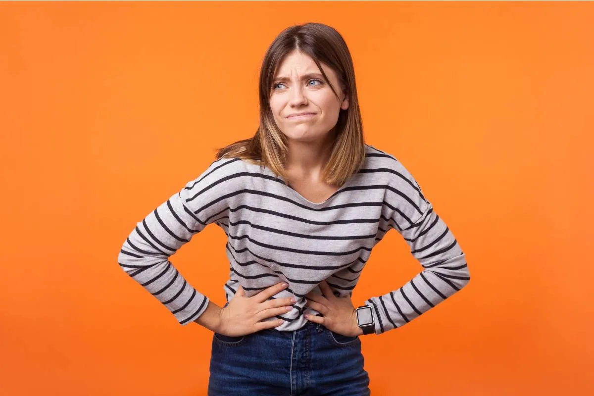 Imagem mostra uma mulher com dor de barriga, Ela está em um fundo laranja usando blusa listrada e calça preta.