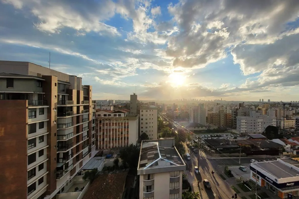 Imagem mostra o céu de Curitiba com prédios e um por do sol entre nuvens.