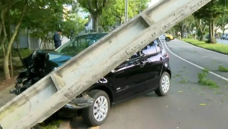 Imagem mostra um carro preto batido contra um poste que está pendendo para a pista.