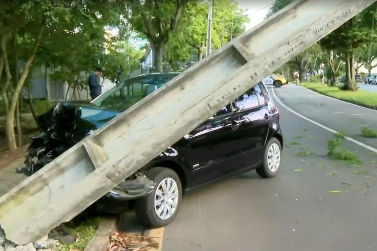 Imagem mostra um carro preto batido contra um poste que está pendendo para a pista.