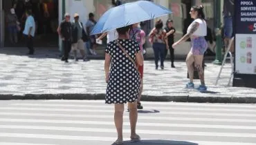 Imagem mostra um dia de muito calo, com uma mulher atravessando a rua com um guarda-chuva escapando do sol escaldante. Há uma mulher com roupa de academia ao fundo e mais pessoas circulando.