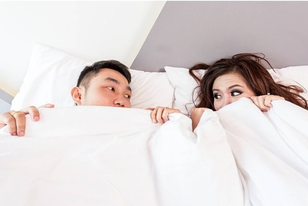 Casal na cama se cobre com uma coberta branca e troca olhares