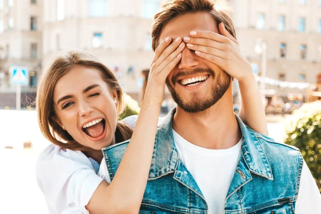 Imagem mostra uma mulher dando risada tampando o olho de um homem com as mãos.