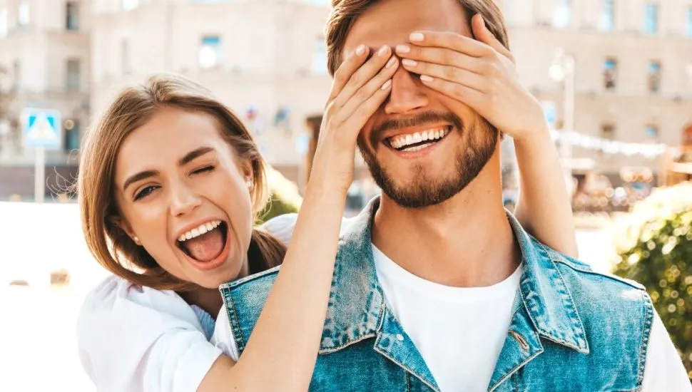 Imagem mostra uma mulher dando risada tampando o olho de um homem com as mãos.