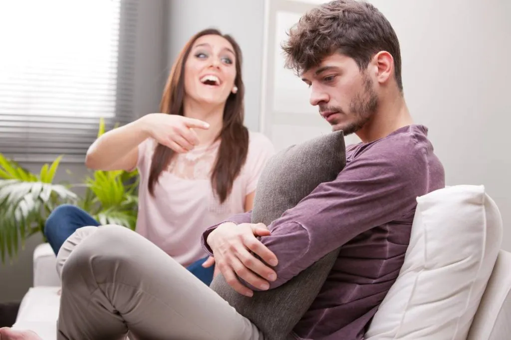 Imagem mostra uma mulher rindo e um homem triste abraçando uma almofada.