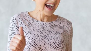 Funny cheerful middle aged blonde woman wearing elegant pencil dress dancing and showing thumbs up gesture as sign of approval, celebrating success or profitable deal, smiling broadly at camera