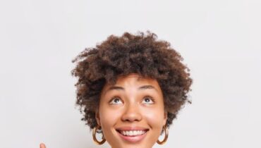 Happy female model with curly hair points up at top advertisement smiles broadly shows special offer or item on discount wears yellow jumper stands against white background. Place your advertisement