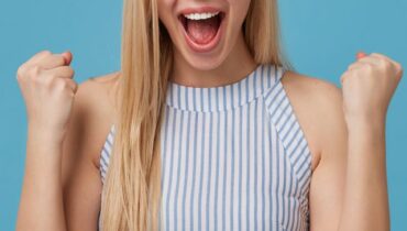 Overjoyed young blonde woman with casual hairstyle raising fists happily, standing over blue background in romantic summer dress, looking at camera with cheerful wide smile