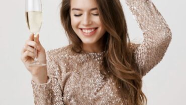 Indoor portrait of joyful attractive caucasian woman dancing with raised glass of champagne, smiling broadly as if partying and celebrating something in trendy evening dress over gray background.