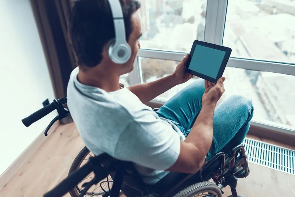 Imagem mostra um jovem sentado numa cadeira de rodas, com fones de ouvido e um tablet nas mãos