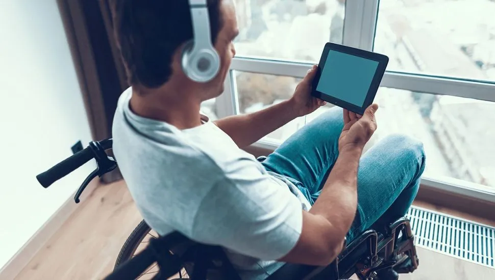 Imagem mostra um jovem sentado numa cadeira de rodas, com fones de ouvido e um tablet nas mãos