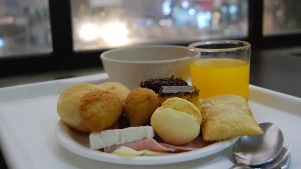 Prato pequeno com coxinha, risoles, pão de queijo, frios, pão, bolo de chocolate, bolo de cenoura, pastel. Copo com suco. Cumbuca com sopa.