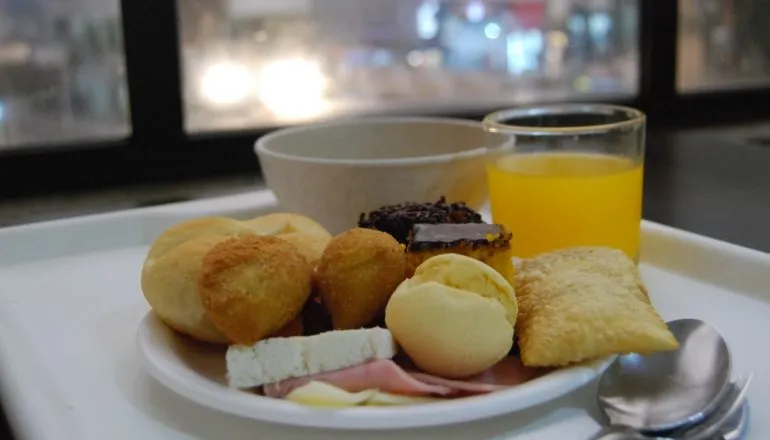 Prato pequeno com coxinha, risoles, pão de queijo, frios, pão, bolo de chocolate, bolo de cenoura, pastel. Copo com suco. Cumbuca com sopa.